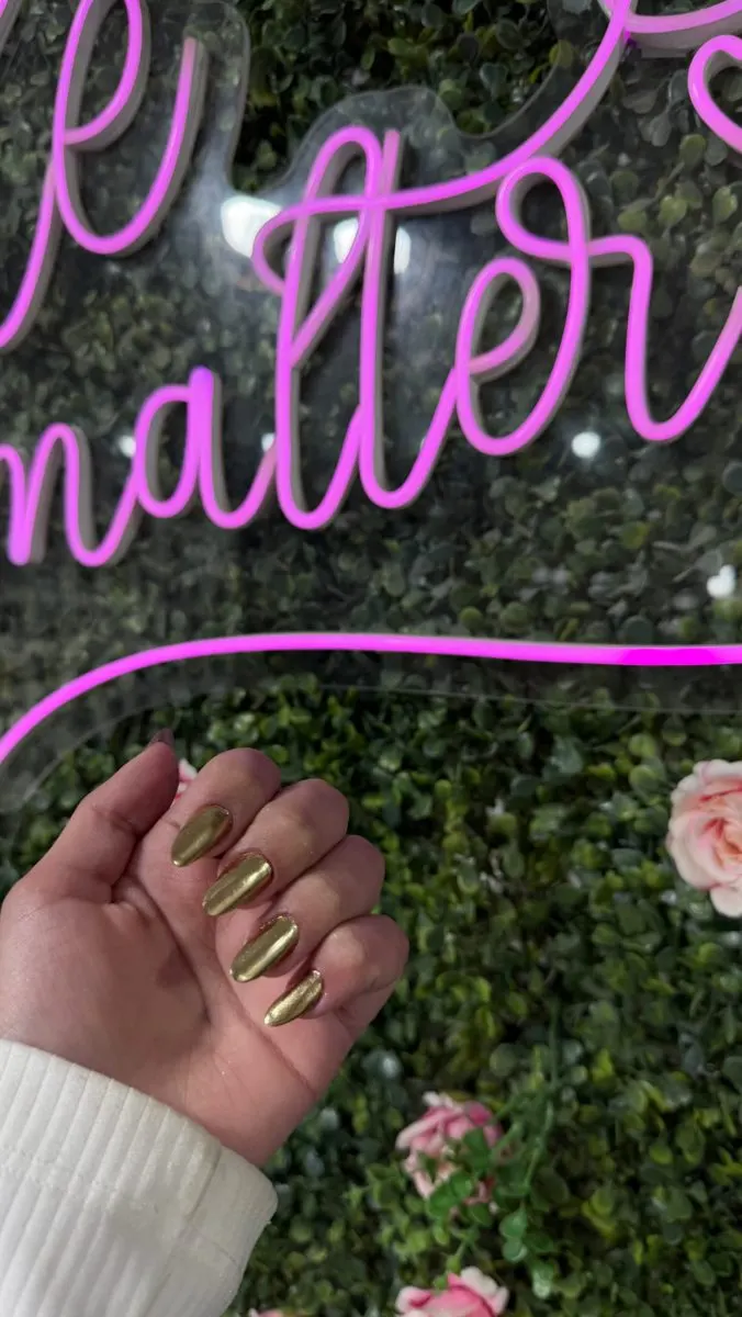 Letreiro neon rosa “Smile Esmalteria” na parede de flores do salão, com uma mão de unhas feitas em primeiro plano